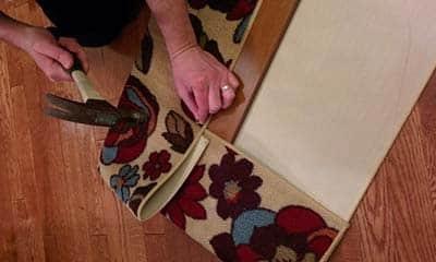 Woman hammering tack nails into board to secure a flowered area rug to wood for cat scratch board.